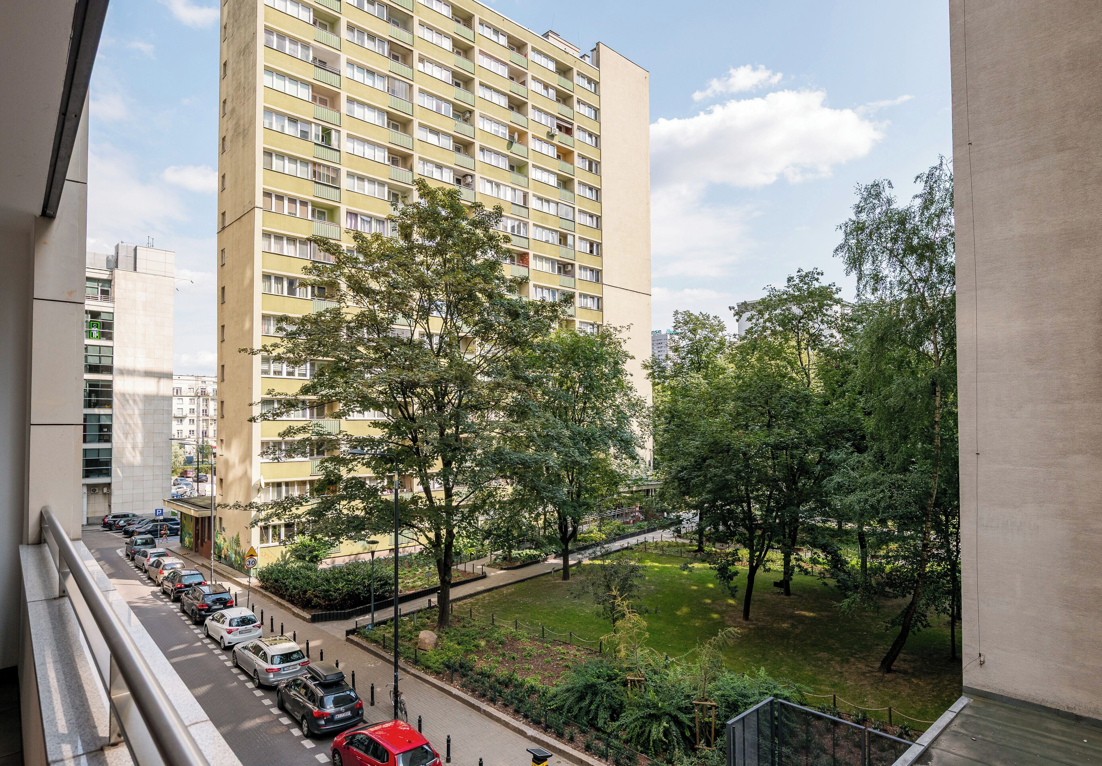 A high-rise building with multiple balconies, a parking lot with cars, and a green park area.