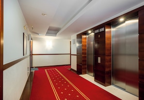 Un couloir d’hôtel avec un tapis rouge, des boiseries et deux ascenseurs.