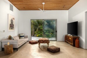 A modern living room with a sofa, ottomans, a television, and a large sliding glass door leading to a tropical garden view.