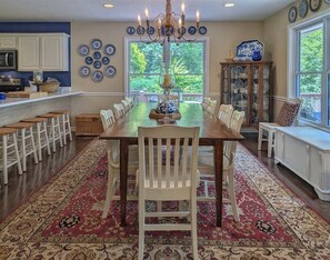 A dining area with a long table, chairs, a chandelier, and a view of greenery outside. - The Haven / Two Adjoining Lake Homes / Blue Ridge Mountain Views (Lake Junaluska)