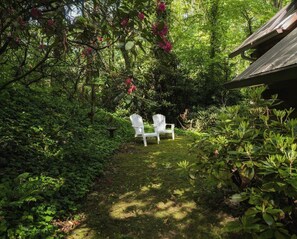 A garden with two white chairs, lush greenery, and a wooden structure. - The Haven / Two Adjoining Lake Homes / Blue Ridge Mountain Views (Lake Junaluska)