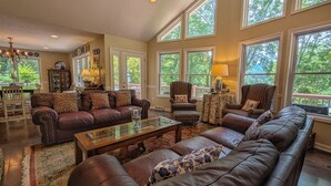 A spacious living room with leather sofas, a glass coffee table, and large windows offering a view of greenery. - The Haven Adjoining houses for large groups/families Lake Junaluska (Lake Junaluska)