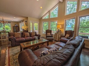 A spacious living room with leather sofas, a glass coffee table, and large windows offering a view of greenery. - The Haven Adjoining houses for large groups/families Lake Junaluska (Lake Junaluska)