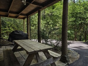 Une table de pique-nique en bois, placée sous un abri et agrémentée d’un barbecue, le tout entouré d’arbres.
