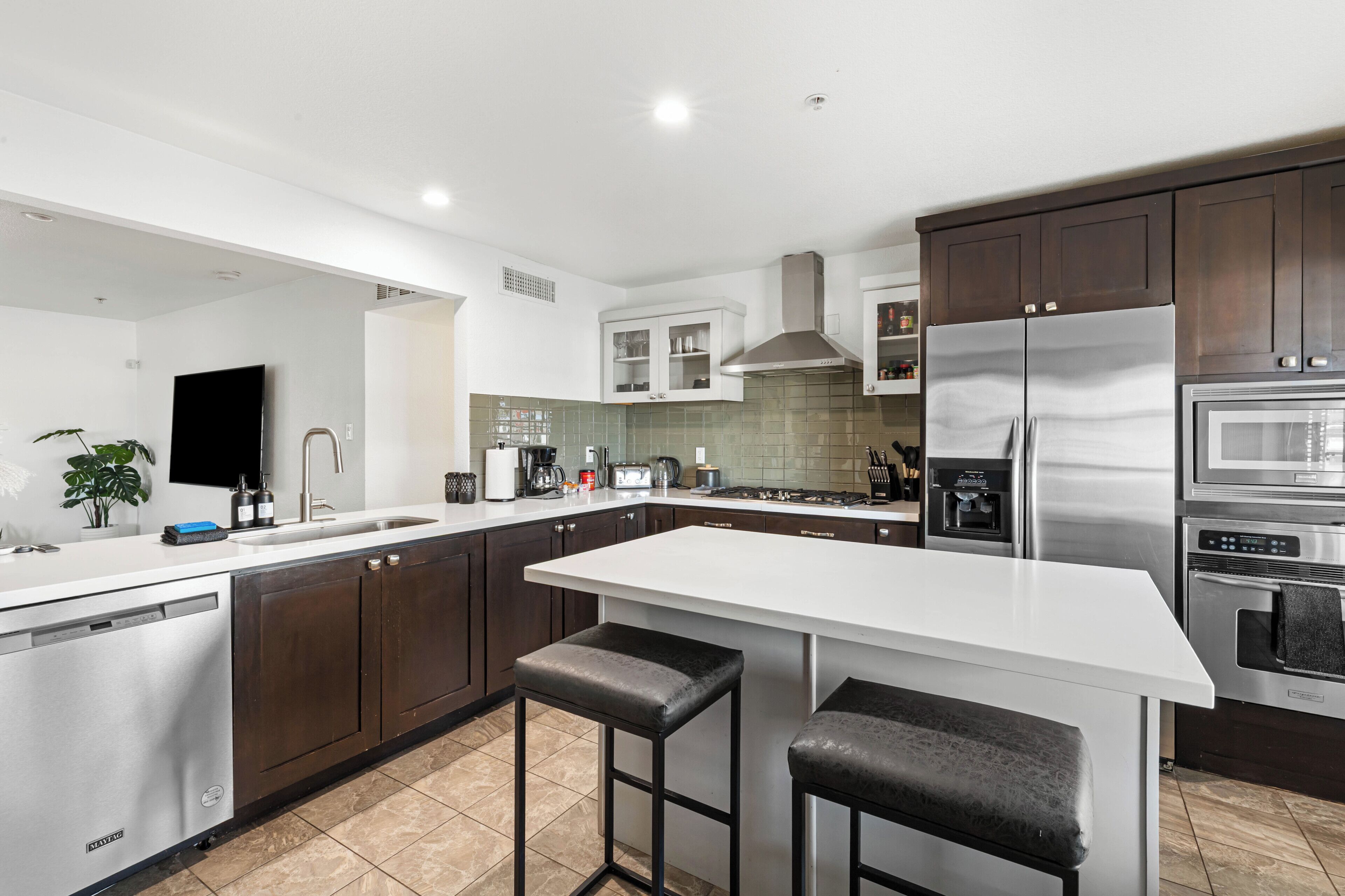 A modern kitchen with dark wooden cabinets, a white countertop, stainless steel appliances, and a bar stool.