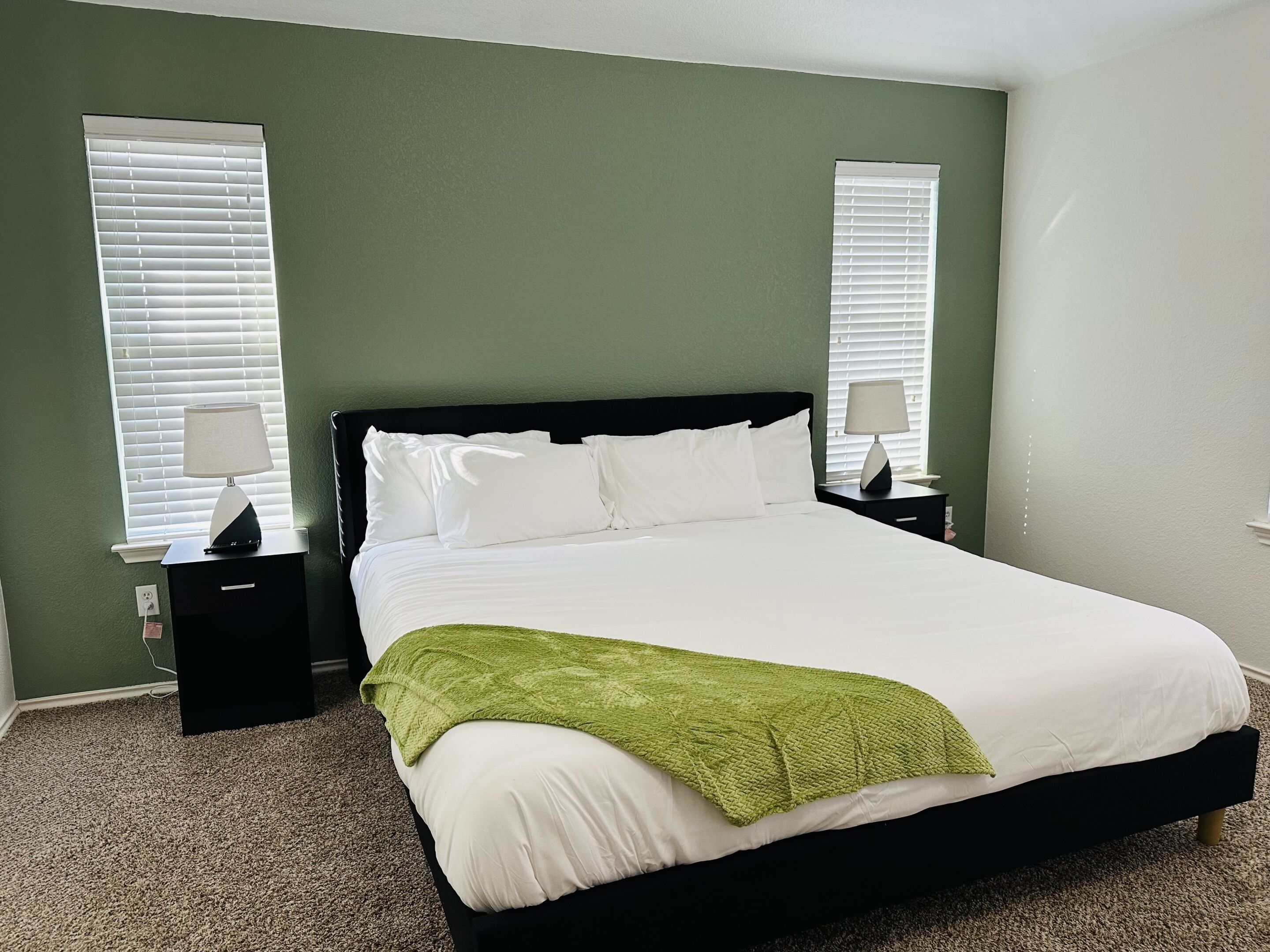 A neatly made bed with white linens and a green throw blanket, flanked by two bedside tables with lamps, in a room with green walls and carpeted floor.