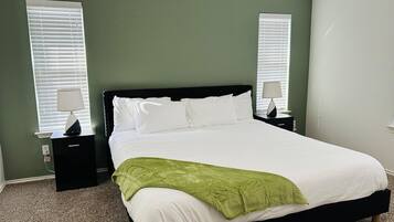 A neatly made bed with white linens and a green throw blanket, flanked by two bedside tables with lamps, in a room with green walls and carpeted floor.