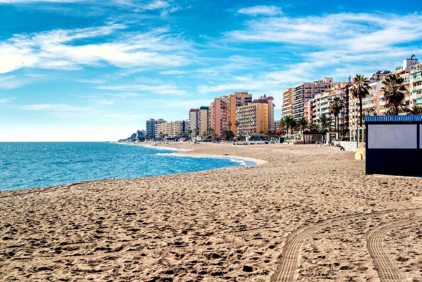 A sandy beach with a clear blue sky, a calm sea, and a row of multi-story buildings in the background.