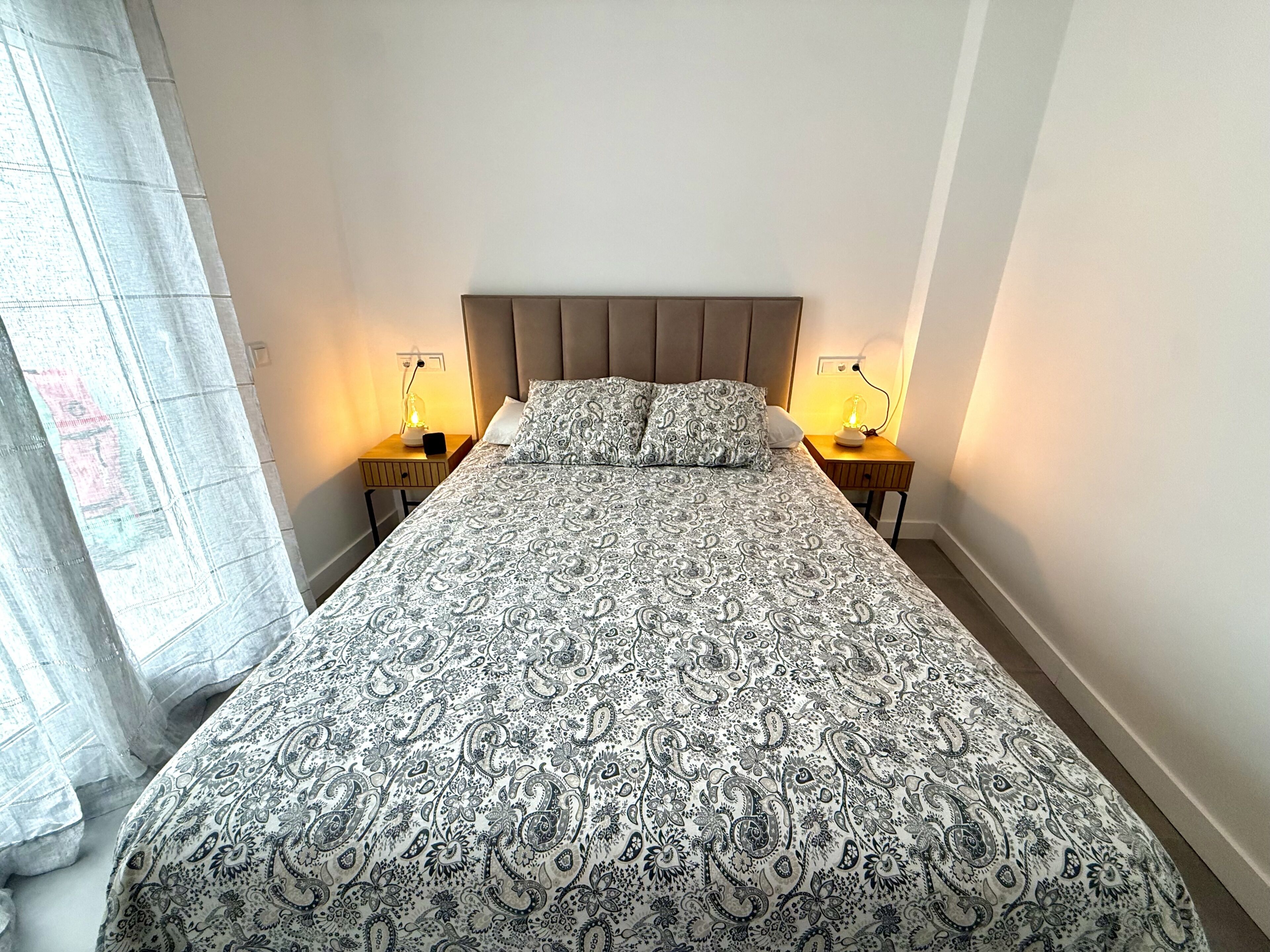 Uma cama com colcha estampada, duas luminárias de cabeceira e uma janela com cortinas leves.
