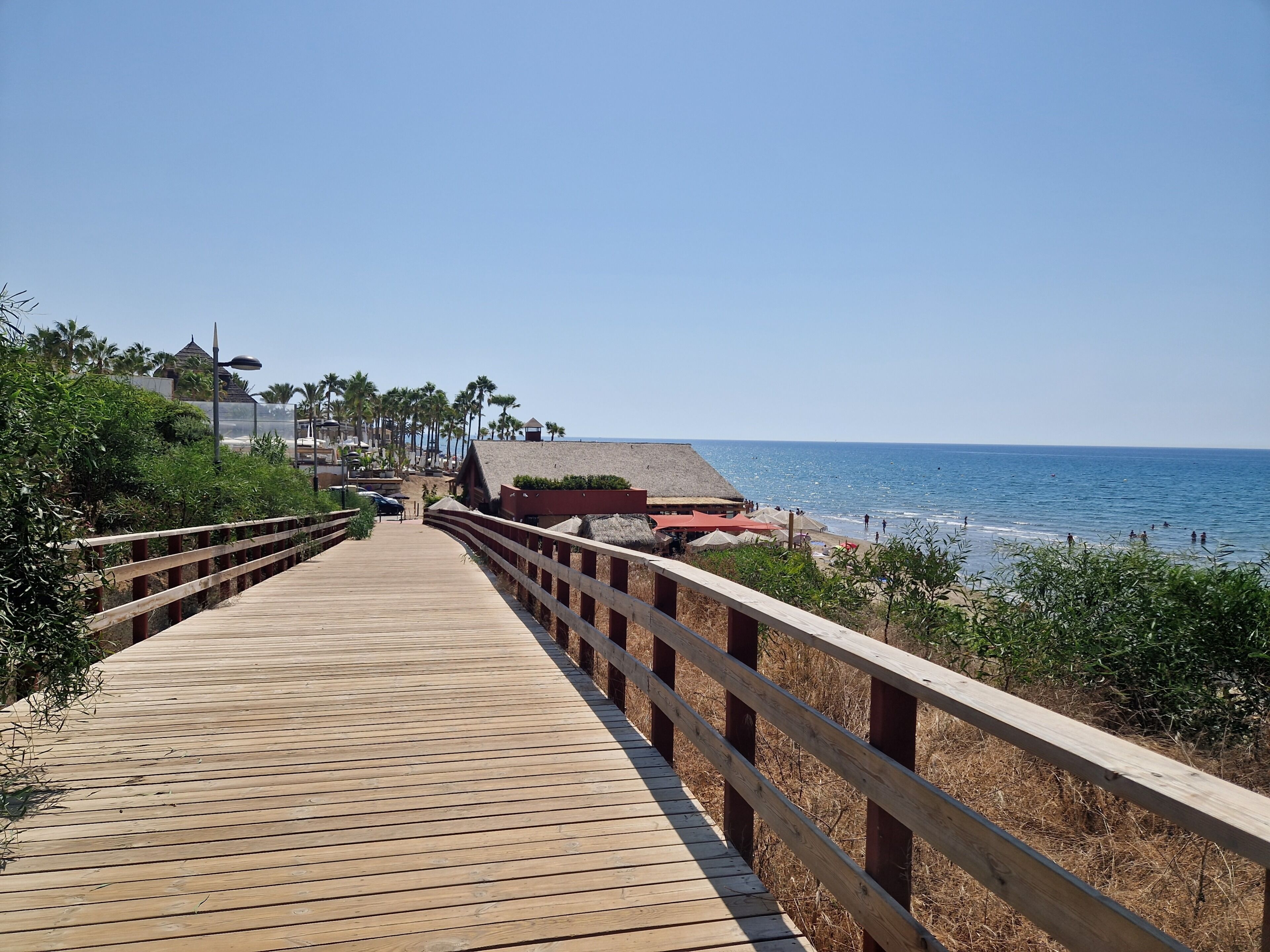 Eine Holzpromenade führt zu einem Strand mit Palmen und strohgedeckten Gebäuden.