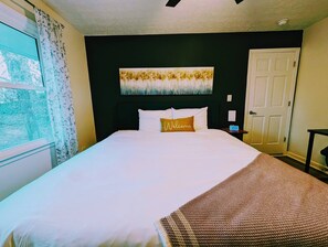 A hotel room with a bed, a painting above the bed, a window with curtains, and a door. - Colony Home for Family Groups - Patio, Workspace (Marietta)