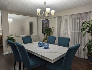 A dining room with a marble table, blue upholstered chairs, a chandelier, and a large window with sheer curtains. - Colony Home for Family Groups - Patio, Workspace (Marietta)