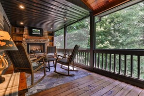 A wooden deck with a fireplace, seating, and a large window overlooking a forest. - Chestnut Hills Lodge by Mountain Oasis | Luxury Blue Ridge Cabin (Blue Ridge)