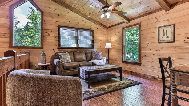 Ein gemütliches Blockhaushaus-Interieur mit Holzbalkendecke, Deckenventilator, einer Couch, einem Couchtisch und einem Esstisch.