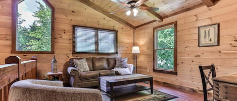 A cozy cabin interior with a wooden ceiling, a ceiling fan, a sofa, a coffee table, and a dining table.
