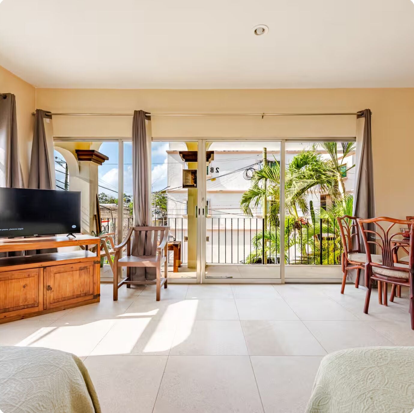 A spacious living area with a television, wooden furniture, and a balcony with a view of palm trees.
