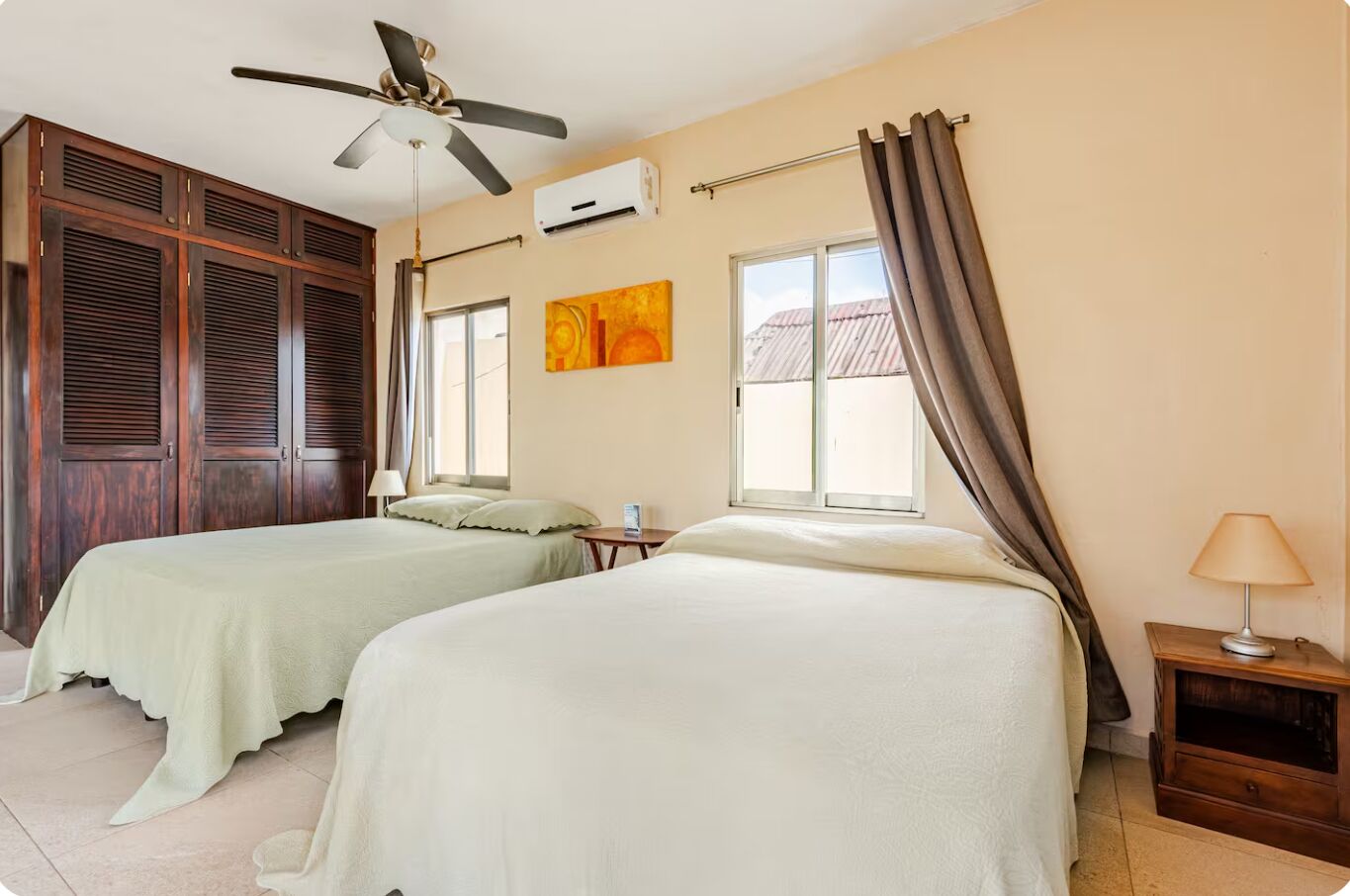 Ein Hotelzimmer mit zwei Betten, einem Deckenventilator, einer Klimaanlage, einem Fenster mit Vorhängen und einem Nachttisch mit einer Lampe.