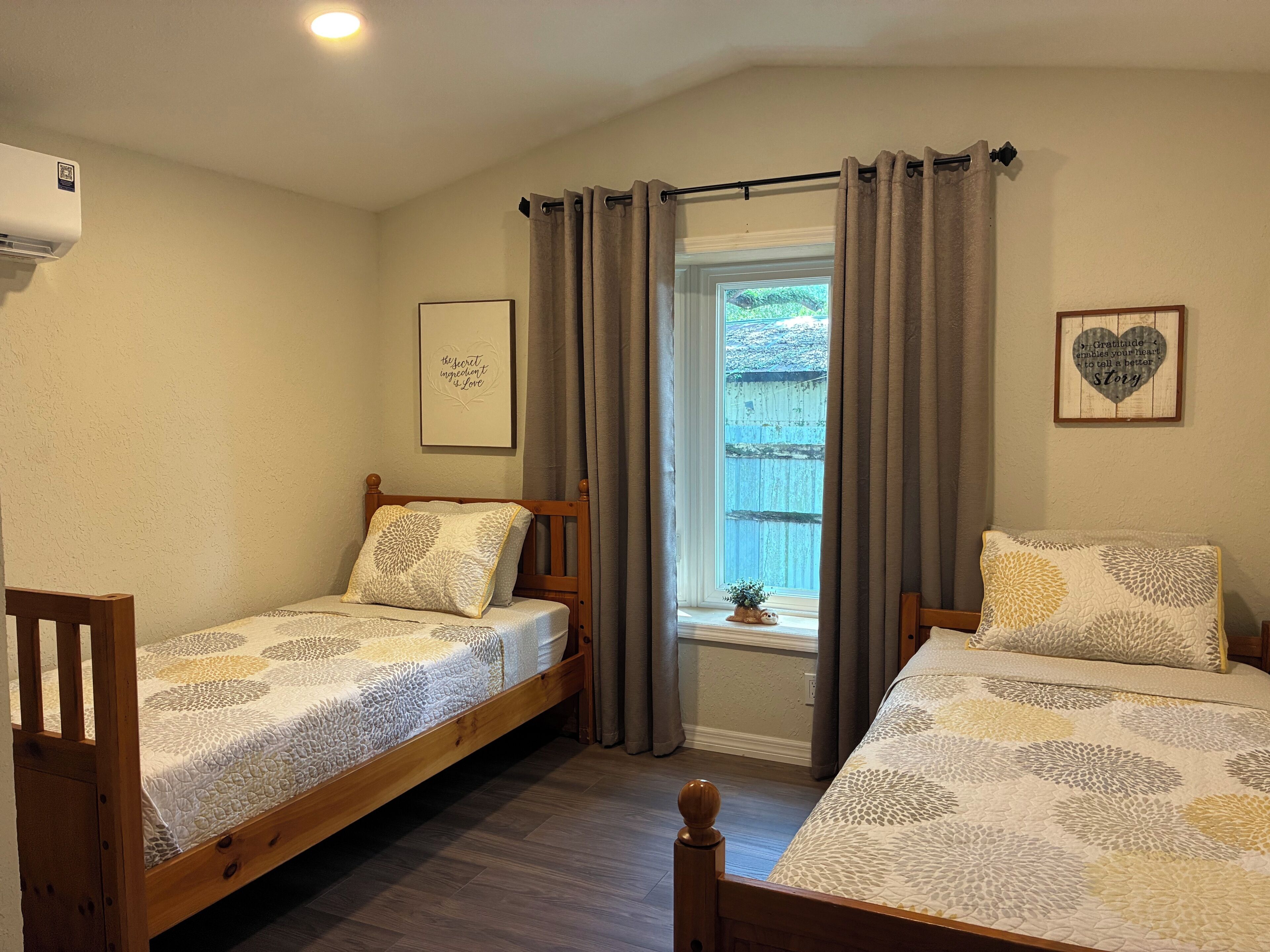 Une chambre à coucher avec deux lits en bois, une fenêtre avec des rideaux, un climatiseur fixé au mur et deux tableaux encadrés.