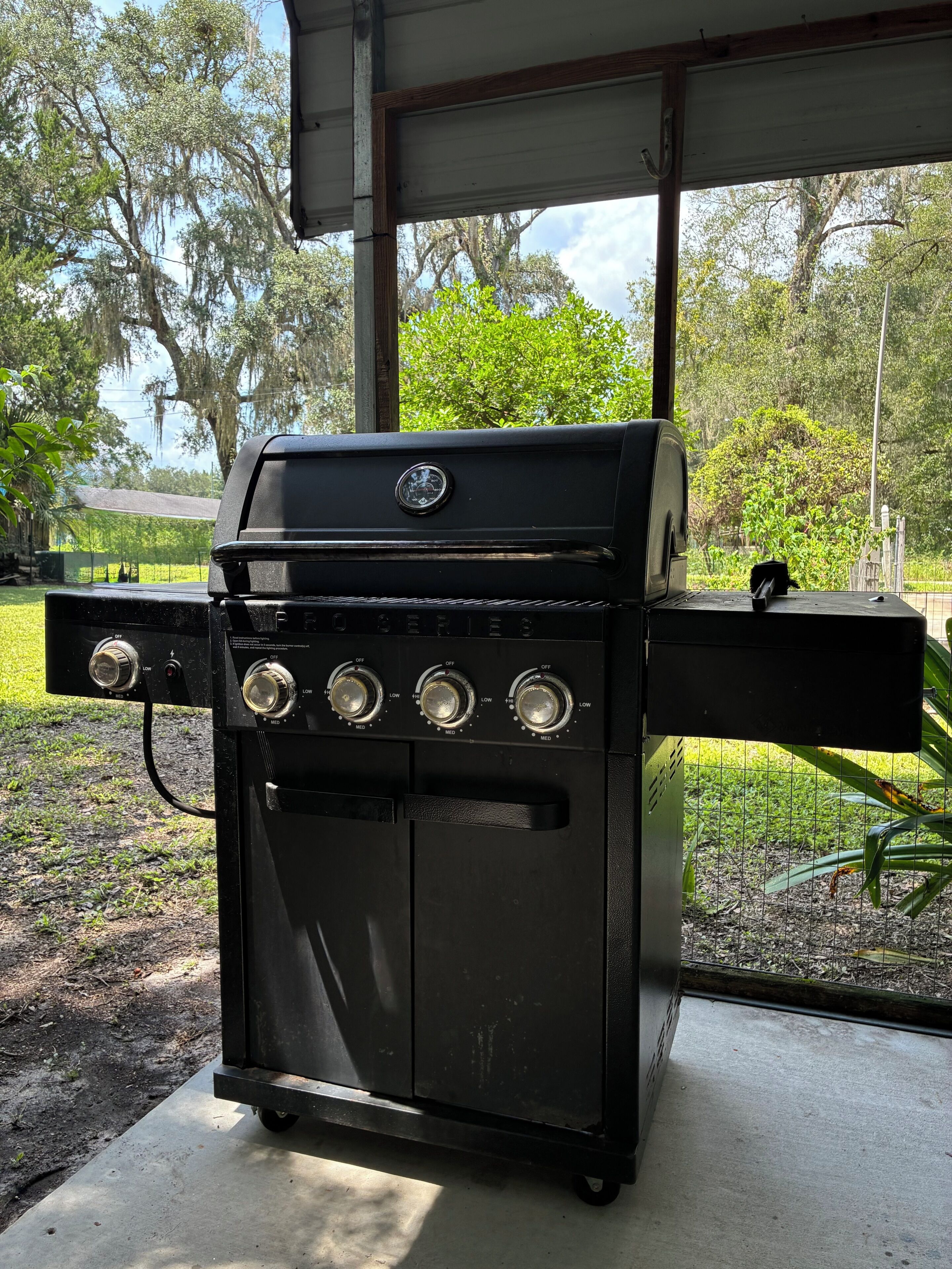 A black barbecue grill with four burners, positioned outdoors with a view of trees and a building in the background.