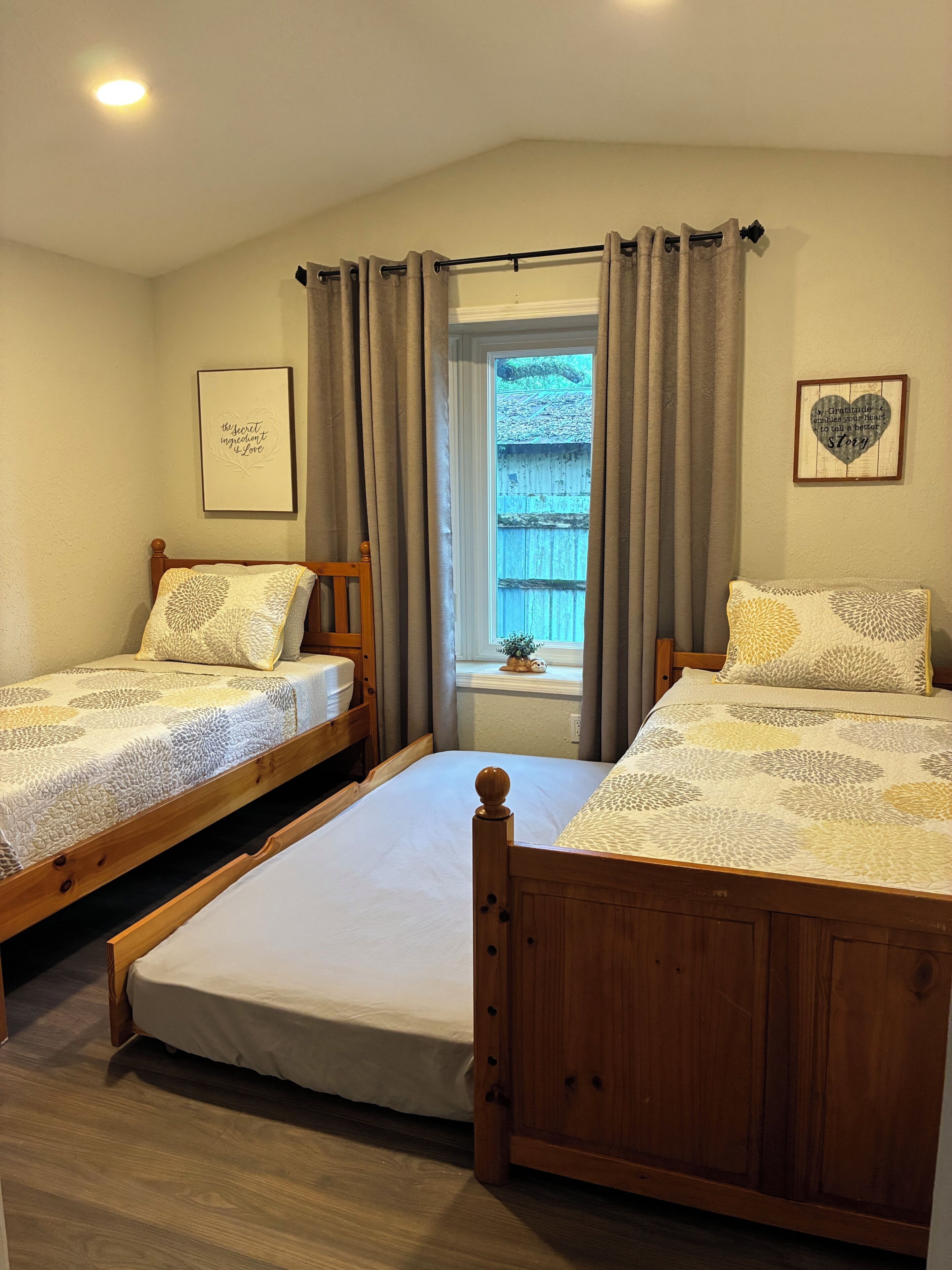 A bedroom with two wooden beds, a window with curtains, a framed quote on the wall, and a small plant on the windowsill.