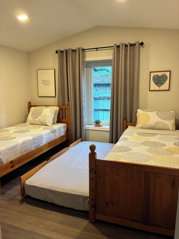 A bedroom with two wooden beds, a window with curtains, a framed quote on the wall, and a small plant on the windowsill.