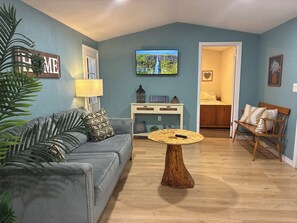 A living room with a grey sofa, a round wooden coffee table, a TV on the wall, and a door leading to another room.