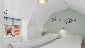 Um quarto com teto inclinado, uma cama com roupa de cama listrada, uma luminária na parede e uma janela com vista para um telhado de telhas.
