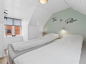 Ein Schlafzimmer mit schrägverdeckter Decke, einem Bett mit gestreifter Bettwäsche, einer Wandlampe und einem Fenster mit Blick auf ein Ziegeldach.