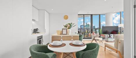 A modern living space with a dining area featuring two green chairs, a round table, and a kitchen with a coffee machine.