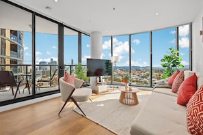 A modern living room with a large TV, comfortable seating, and a view of a cityscape through floor-to-ceiling windows.