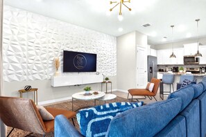 Un salon moderne avec un canapé d’angle, un téléviseur à écran plat fixé sur un mur orné d’un motif géométrique, un coin cuisine doté d’appareils électroménagers en acier inoxydable et un espace repas avec des tabourets de bar.