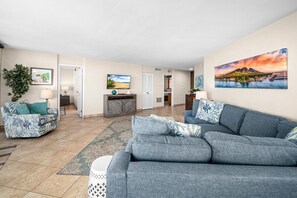 Ein geräumiges Wohnzimmer mit grauem Sofa, einem gemusterten Teppich und einem Fernseher auf einer Holzkommode.