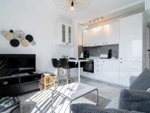 Una sala moderna con un televisor de pantalla plana, una mesa de centro y un sofá. Cuenta con una zona de cocina con armarios blancos y una mesa de comedor con sillas.