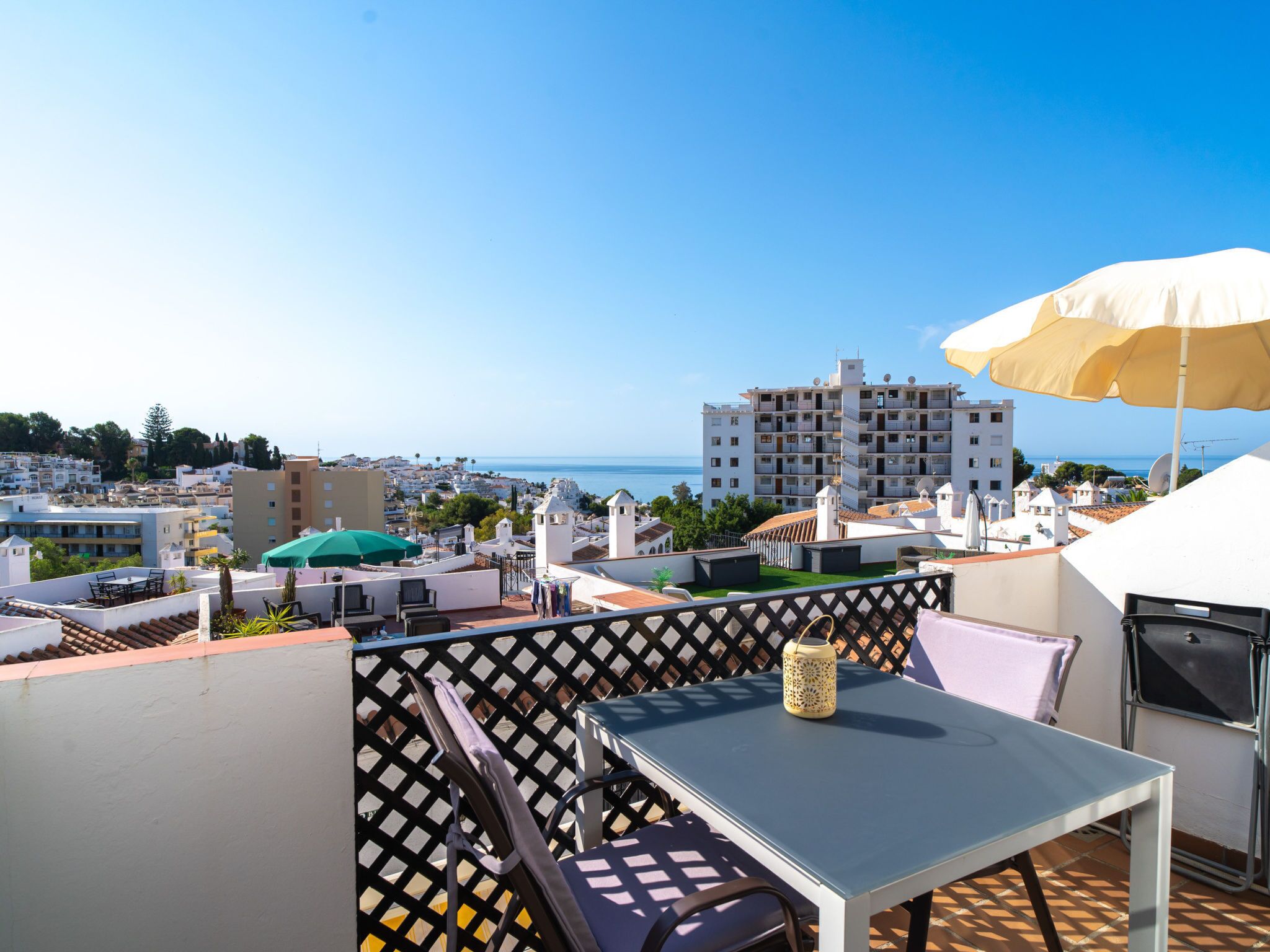 Una terraza en la azotea con una mesa, sillas, una sombrilla grande y vistas al mar y edificios.