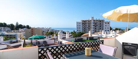 A rooftop terrace with a table and chairs, a large umbrella, and a view of the sea and buildings.