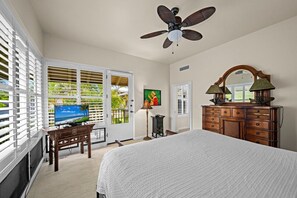Ein Schlafzimmer mit einem Bett, einem Deckenventilator, einem Schminktisch, einem Fernseher und einer Tür, die auf einen Balkon mit Blick auf Palmen führt.