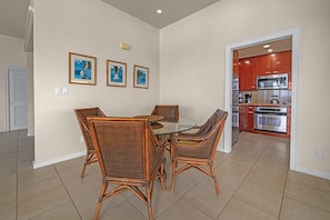 A dining area with a round table and four wicker chairs, a kitchen with red cabinets and stainless steel appliances, and three framed pictures on the wall. - Kapalua Bay Villa 22B4 Gold Oceanfront 1 Bedroom (Lahaina)