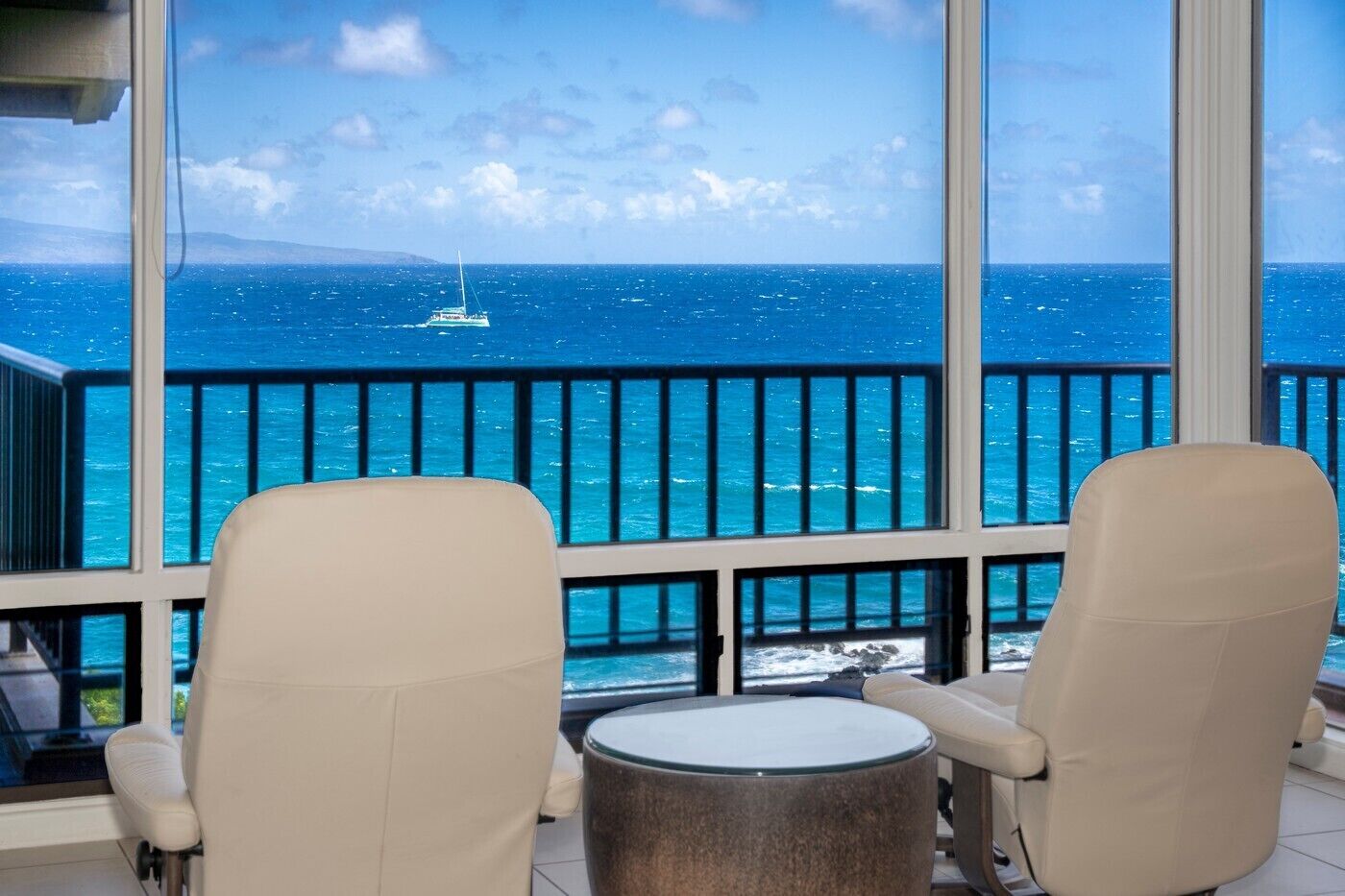Un balcon avec deux chaises beiges et une petite table ronde, donnant sur une mer bleue où l’on aperçoit un voilier.