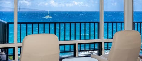 A balcony with two beige chairs and a small round table, overlooking a blue sea with a sailboat.