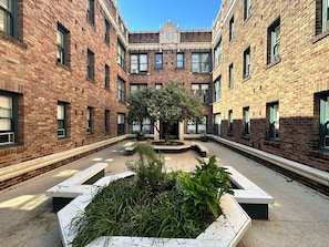A courtyard with brick buildings, a central tree, and planters with greenery.