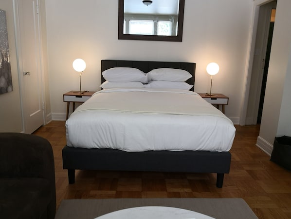 A bedroom with a large bed, two bedside tables with lamps, a mirror above the bed, and a door to the left.