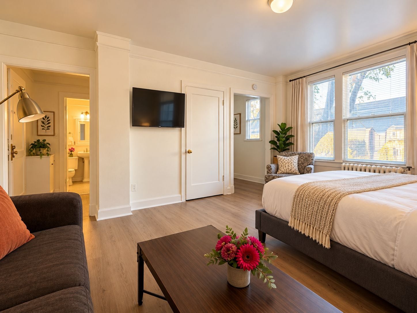 A bedroom with a bed, a sofa, a coffee table with flowers, a TV, and a window with blinds.