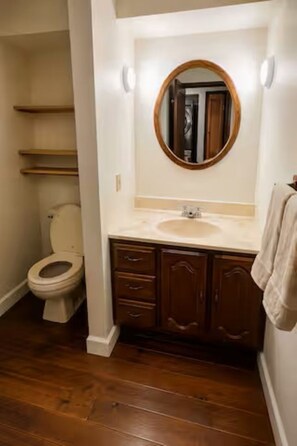 A bathroom with a toilet, a sink with a mirror, wooden cabinets, and a wooden floor. - Lakefront 3-Bedroom Retreat on Huffman Lake (Elmira)