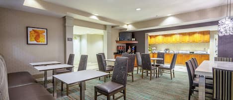 A hotel dining area with tables, chairs, a sofa, a kitchen in the background, and a framed picture on the wall.