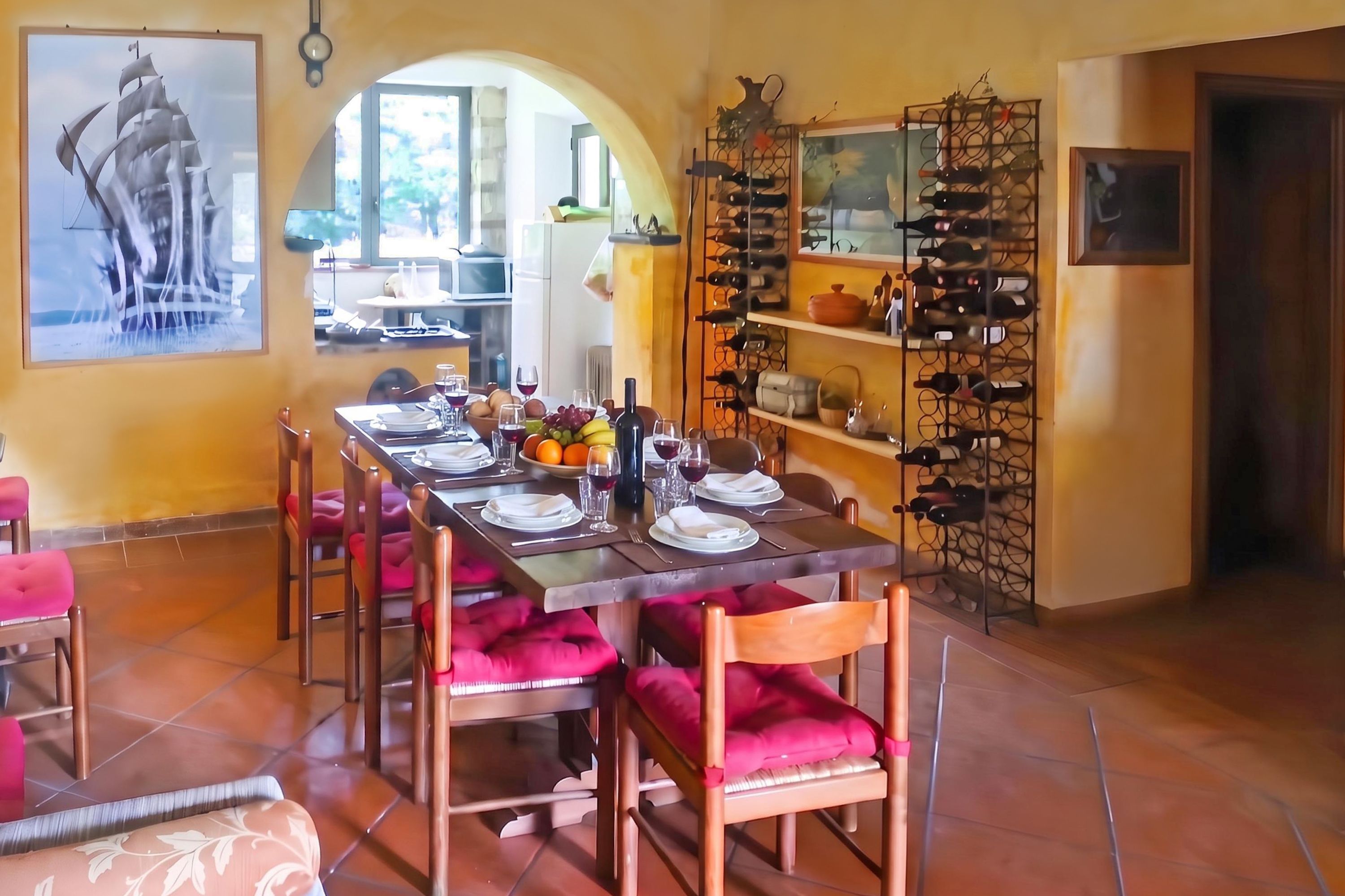 Un coin repas avec une longue table dressée pour quatre personnes, entourée de chaises dotées de coussins rouges. Un meuble à bouteilles est fixé au mur, à droite.