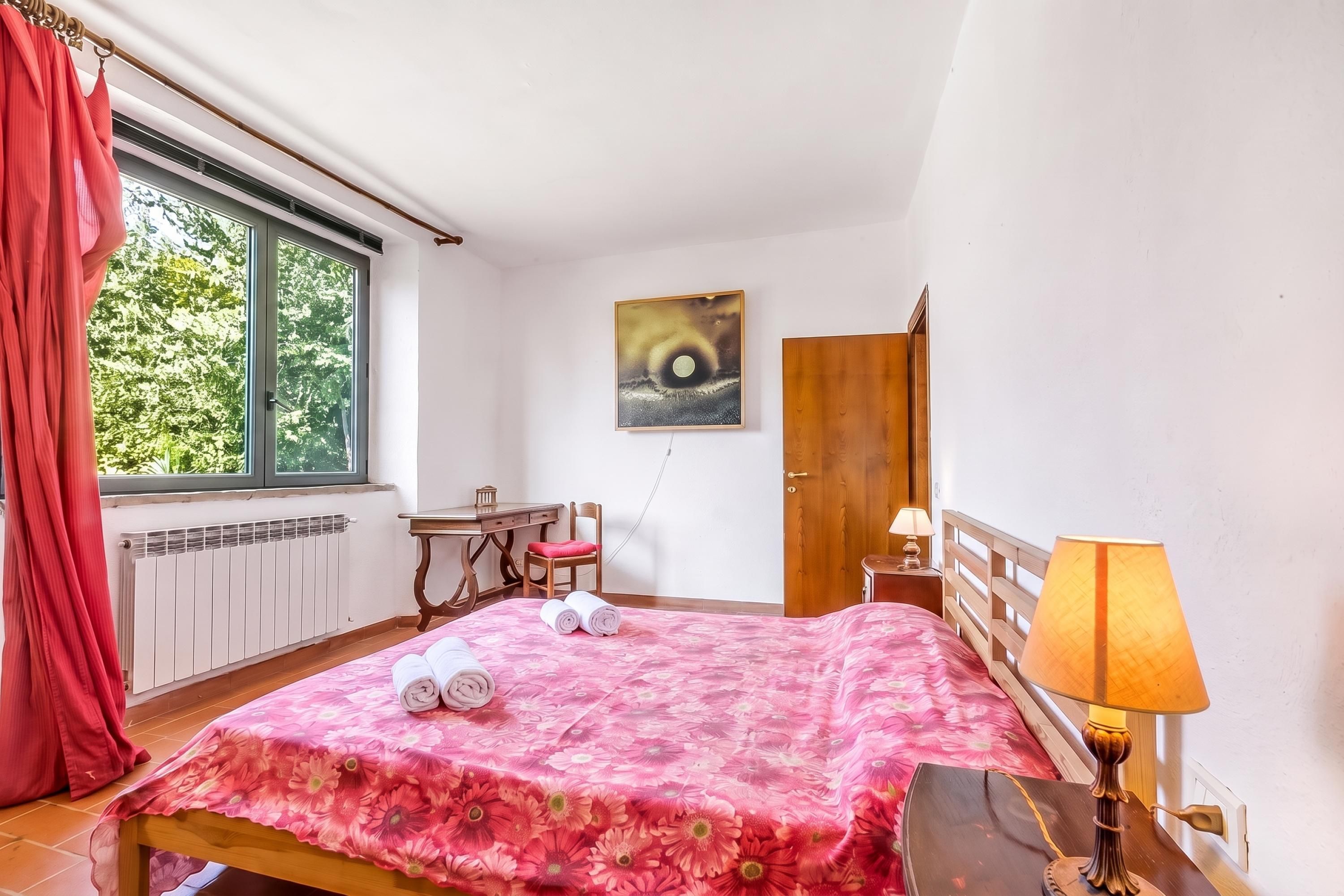 Ein Schlafzimmer mit einem Bett, einem Schreibtisch, einem Stuhl, einem Fenster mit Vorhängen, einem Bild an der Wand und einer Tür.