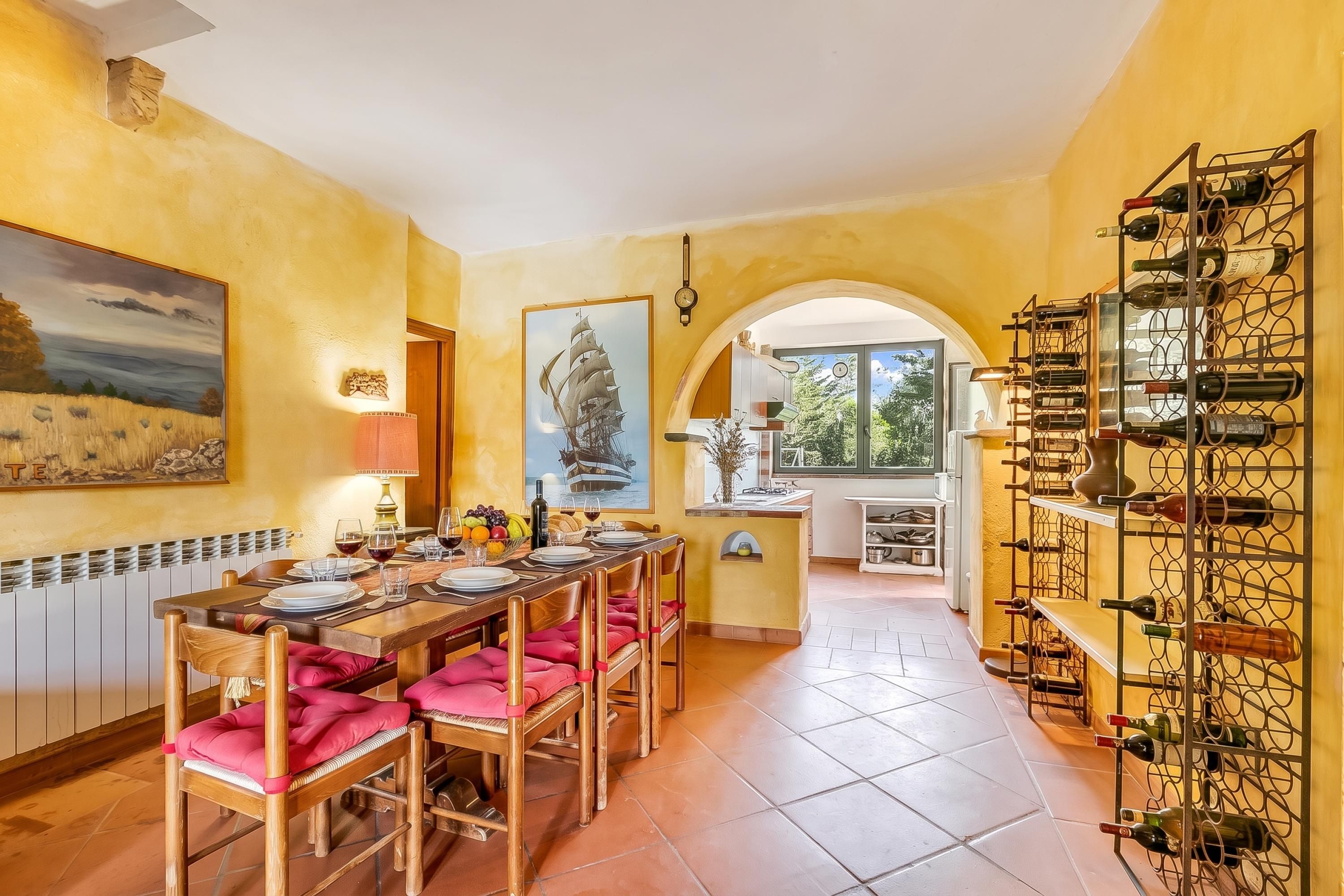 Un coin repas avec une longue table dressée pour quatre personnes, un meuble à vin sur le côté droit et une vue sur la cuisine à travers une arche.
