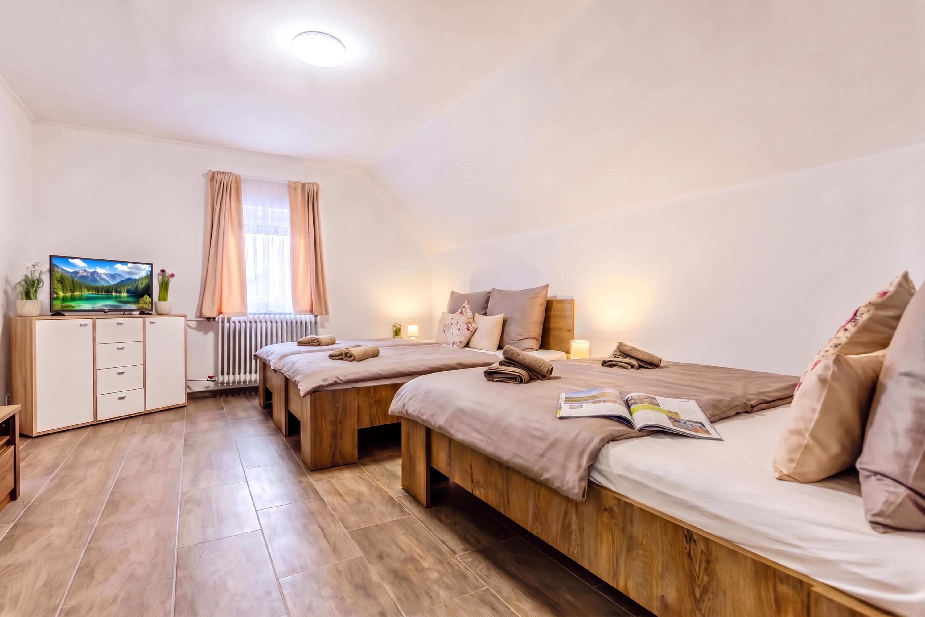 A hotel room with two beds, a wooden floor, a radiator, a TV on a cabinet, and a window with curtains.