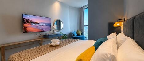 Quarto de hotel moderno com cama, televisão de tela plana, mesa de cabeceira, espelho e janela com vista.