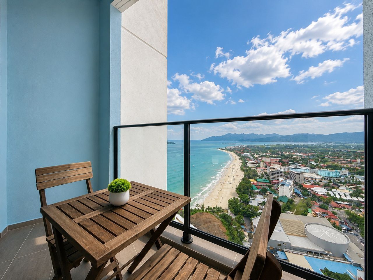 A balcony with a wooden table and chairs, a potted plant, and a view of a beach and cityscape.